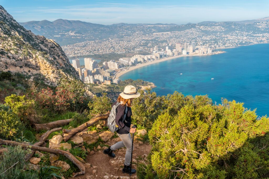 Ein junger Wanderer mit Hut auf dem Abstiegspfad des Penon de Ifach Naturparks mit der Stadt Calpe im Hintergrund, Valencia. Spain. Das mediterrane Meer. Blick auf den Strand von La Fossa