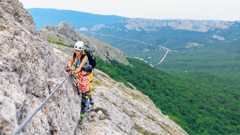 Junge Mutter, Kind in Sicherheitsausrüstung, Aufstieg zum Gipfel über die Klettersteigroute für Anfänger. Familienreiseabenteuer, Hiking-Aktivität. Kinder erkunden die Natur während eines Sommerurlaubs. Wochenendtägige Wandertour